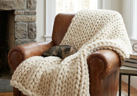 A tabby cat sleeps on a chunky cream knit blanket draped over a brown leather armchair in a cozy living room